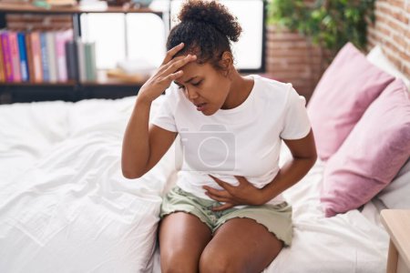 Téléchargez les photos : Femme afro-américaine souffrant de maux d'estomac et de tête assis sur le lit dans la chambre - en image libre de droit