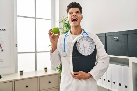 Foto de Joven médico hispano sosteniendo balanza en clínica dietista enojado y loco gritando frustrado y furioso, gritando con rabia mirando hacia arriba. - Imagen libre de derechos