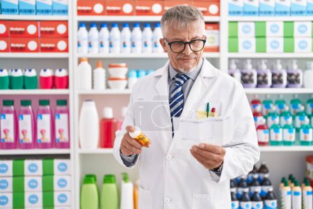 Téléchargez les photos : Homme d'âge moyen aux cheveux gris pharmacien tenant des pilules bouteille lecture prescription au laboratoire - en image libre de droit