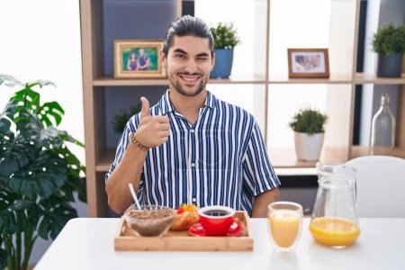 Foto de Hombre hispano con el pelo largo sentado en la mesa desayunando sonriendo feliz y positivo, pulgar hacia arriba haciendo excelente y signo de aprobación - Imagen libre de derechos