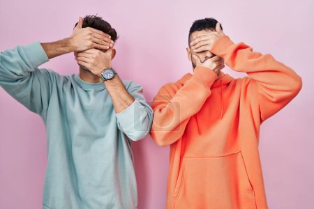 Foto de Joven pareja gay hispana de pie sobre fondo rosa cubriendo los ojos y la boca con las manos, sorprendida y conmocionada. ocultar emociones - Imagen libre de derechos