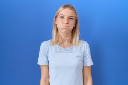 Foto de Mujer caucásica joven que usa casual camiseta azul soplando mejillas con la cara divertida. boca hinchada de aire, expresión loca. - Imagen libre de derechos