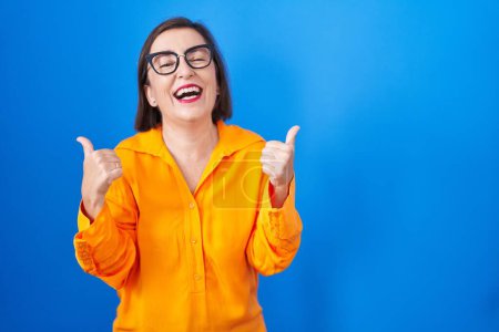 Téléchargez les photos : Femme hispanique du Moyen Age portant des lunettes debout sur fond bleu signe de succès faire un geste positif avec la main, pouces levés souriant et heureux. expression joyeuse et geste gagnant. - en image libre de droit