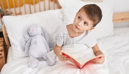 Photo for Adorable caucasian boy reading book sitting on bed at bedroom - Royalty Free Image