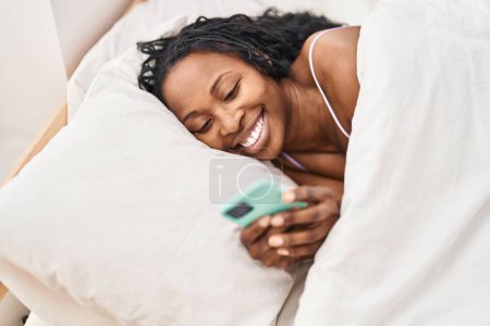 Photo for African american woman using smartphone lying on bed at bedroom - Royalty Free Image