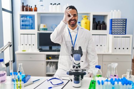 Foto de Joven hispano con barba trabajando en el laboratorio científico haciendo un buen gesto con la mano sonriente, los ojos mirando a través de los dedos con cara feliz. - Imagen libre de derechos