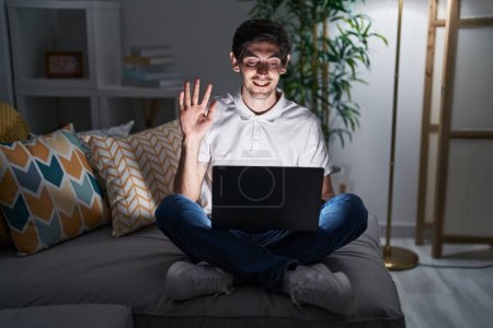 Foto de Joven hombre hispano usando portátil en casa por la noche renunciando a decir hola feliz y sonriente, gesto de bienvenida amistoso - Imagen libre de derechos