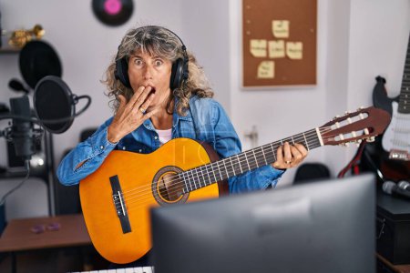 Foto de Mujer de mediana edad tocando la guitarra clásica en el estudio de música cubriendo la boca con la mano, sorprendido y asustado por error. expresión sorprendida - Imagen libre de derechos