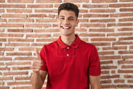 Photo for Young hispanic man standing over bricks wall doing happy thumbs up gesture with hand. approving expression looking at the camera showing success. - Royalty Free Image