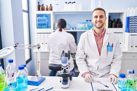 Foto de Hombre joven que trabaja en el laboratorio científico mirando positivo y feliz de pie y sonriendo con una sonrisa confiada mostrando los dientes - Imagen libre de derechos