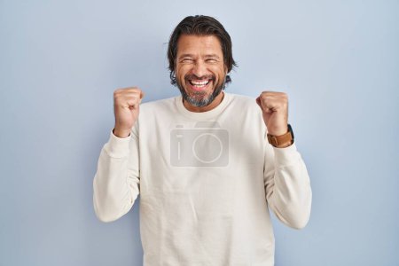 Foto de Hombre guapo de mediana edad con suéter casual sobre fondo azul emocionado por el éxito con los brazos levantados y los ojos cerrados celebrando la victoria sonriendo. concepto ganador. - Imagen libre de derechos