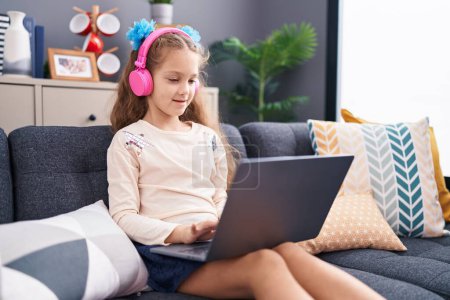 Photo for Adorable caucasian girl using laptop and headphones sitting on sofa at home - Royalty Free Image