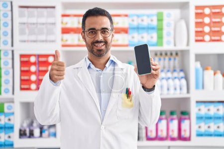 Photo for Hispanic man with beard working at pharmacy drugstore showing smartphone screen smiling happy and positive, thumb up doing excellent and approval sign - Royalty Free Image