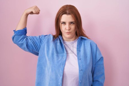 Foto de Mujer hispana joven con el pelo rojo de pie sobre el fondo rosa persona fuerte mostrando el músculo del brazo, confiado y orgulloso de poder - Imagen libre de derechos