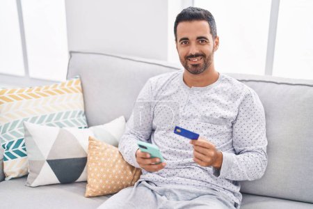 Photo for Young hispanic man using smartphone and credit card sitting on sofa at home - Royalty Free Image
