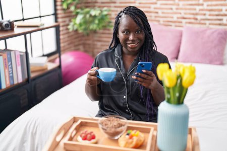 Foto de Mujer afroamericana desayunando con un smartphone en el dormitorio - Imagen libre de derechos