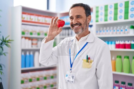Téléchargez les photos : Homme moyen pharmacien souriant confiant tenant le cœur à la pharmacie - en image libre de droit