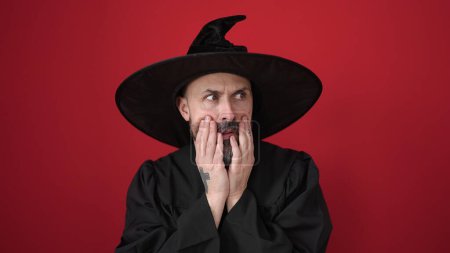 Téléchargez les photos : Jeune homme chauve portant un costume de sorcier debout avec l'expression de la peur sur fond rouge isolé - en image libre de droit