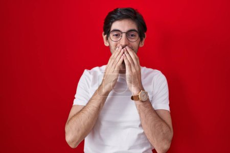 Foto de Young hispanic man standing over red background laughing and embarrassed giggle covering mouth with hands, gossip and scandal concept - Imagen libre de derechos