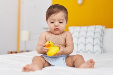 Foto de Adorable bebé caucásico sentado en la cama jugando con el juguete del pato en el dormitorio - Imagen libre de derechos