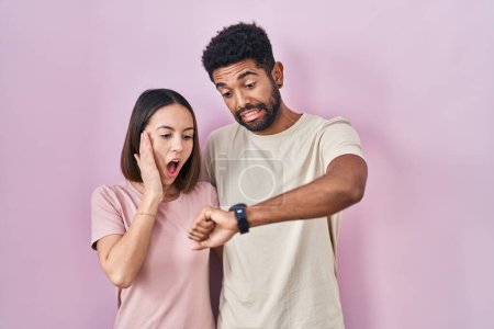 Téléchargez les photos : Jeune couple hispanique ensemble sur fond rose regardant le temps de la montre inquiet, peur de se faire tard - en image libre de droit