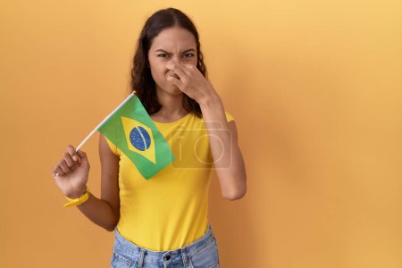 Téléchargez les photos : Jeune femme hispanique tenant drapeau brésilien sentant quelque chose de puant et dégoûtant, odeur intolérable, retenant souffle avec les doigts sur le nez. mauvaise odeur - en image libre de droit