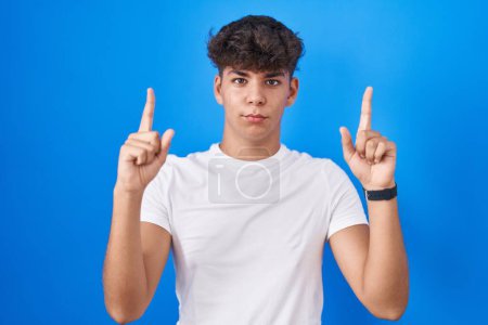 Téléchargez les photos : Adolescent hispanique debout sur fond bleu pointant vers le haut regardant triste et contrarié, indiquant la direction avec les doigts, malheureux et déprimé. - en image libre de droit