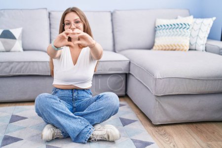 Photo for Young caucasian woman sitting on the floor at the living room smiling in love doing heart symbol shape with hands. romantic concept. - Royalty Free Image