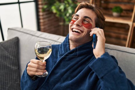 Foto de Joven hombre caucásico usando parches bajo los ojos hablando en el teléfono inteligente en casa - Imagen libre de derechos