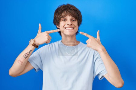 Téléchargez les photos : Jeune homme hispanique debout sur fond bleu souriant gai montrant et pointant avec les doigts dents et la bouche. concept de santé dentaire. - en image libre de droit