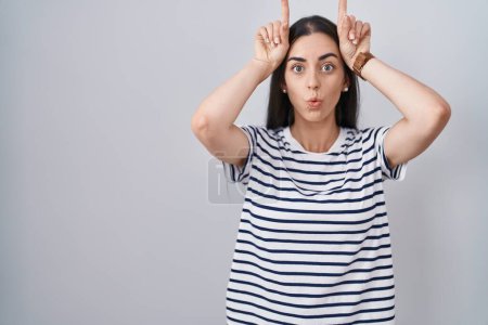 Téléchargez les photos : Jeune femme brune portant un t-shirt rayé faisant un geste drôle avec le doigt sur la tête comme des cornes de taureau - en image libre de droit