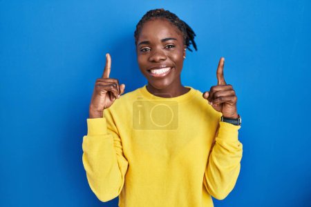Téléchargez les photos : Belle femme noire debout sur fond bleu souriant étonné et surpris et pointant vers le haut avec les doigts et les bras levés. - en image libre de droit