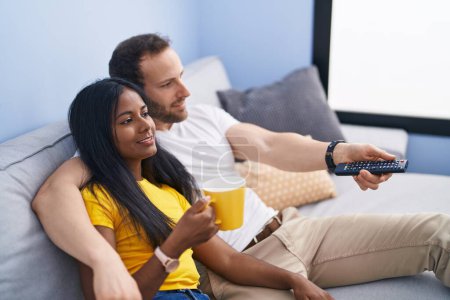 Foto de Hombre y mujer interracial pareja bebiendo café viendo tv en casa - Imagen libre de derechos
