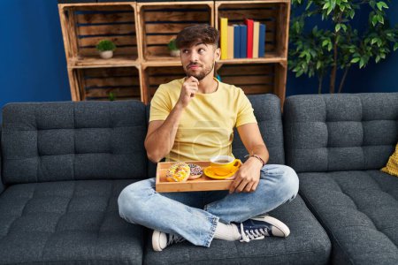 Téléchargez les photos : Homme arabe avec barbe assis sur le canapé à la maison manger le petit déjeuner visage sérieux penser à la question avec la main sur le menton, réfléchi sur l'idée déroutante - en image libre de droit