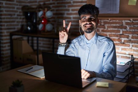 Foto de Joven hombre hispano con barba trabajando en la oficina por la noche mostrando y señalando con los dedos el número dos mientras sonríe confiado y feliz. - Imagen libre de derechos