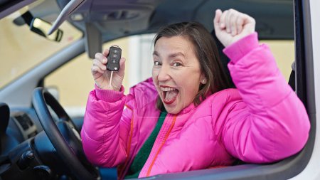 Téléchargez les photos : Femme hispanique mature avec les cheveux gris souriant confiance tenant la clé de la nouvelle voiture à la rue - en image libre de droit