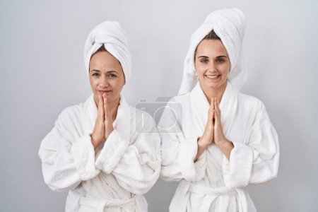 Téléchargez les photos : Femme et fille d'âge moyen portant un peignoir blanc et une serviette priant avec les mains jointes demandant pardon en souriant confiant. - en image libre de droit