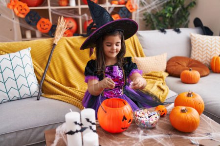 Photo for Adorable hispanic girl having halloween party putting sweet in pumpkin basket at home - Royalty Free Image