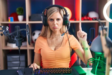 Foto de Mujer joven jugando videojuegos apuntando hacia arriba buscando triste y molesto, indicando la dirección con los dedos, infeliz y deprimido. - Imagen libre de derechos