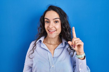 Téléchargez les photos : Jeune femme brune debout sur fond bleu pointant du doigt avec une idée réussie. Sorti et heureux. numéro un. - en image libre de droit