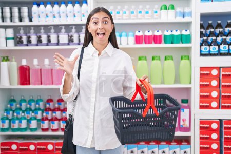 Téléchargez les photos : Jeune femme hispanique faisant du shopping à la pharmacie drugstore tenant panier célébrant la victoire avec sourire heureux et expression gagnante avec les mains levées - en image libre de droit