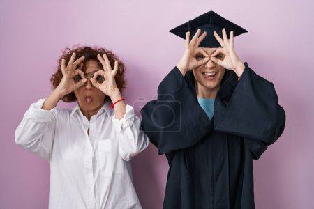 Foto de Madre e hija hispanas usando gorra de graduación y bata de ceremonia haciendo un gesto aceptable como binoculares sacando la lengua, ojos mirando a través de los dedos. expresión loca. - Imagen libre de derechos