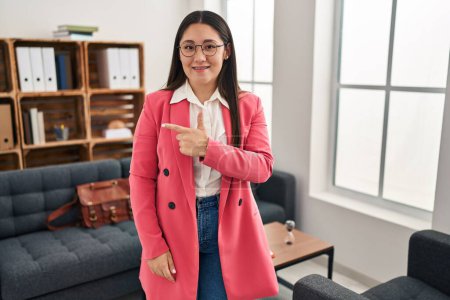 Foto de Mujer latina joven trabajando en la oficina de consulta sonriendo alegre señalando con la mano y el dedo hacia un lado - Imagen libre de derechos