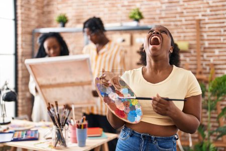 Téléchargez les photos : Jeune peintre noire au studio d'art tenant la palette en colère et folle hurlant frustrée et furieuse, criant de colère en levant les yeux. - en image libre de droit