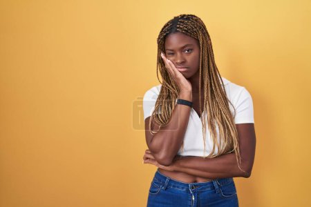 Foto de Mujer afroamericana con cabello trenzado de pie sobre fondo amarillo pensando que se ve cansada y aburrida con problemas de depresión con los brazos cruzados. - Imagen libre de derechos