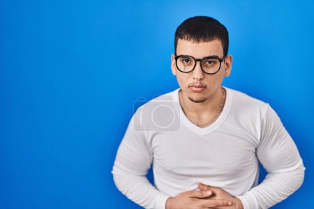 Téléchargez les photos : Jeune homme arabe portant une chemise blanche décontractée et des lunettes avec la main sur le ventre parce que l'indigestion, la maladie douloureuse se sentent mal. concept de douleur. - en image libre de droit