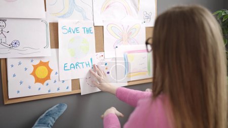 Téléchargez les photos : Jeune femme blonde travaillant comme professeur suspendu sauver la terre dessiner sur le panneau de liège à la maternelle - en image libre de droit