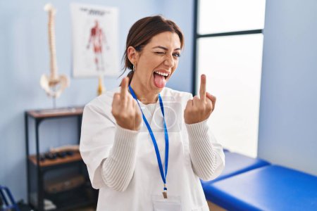 Foto de Young brunette woman working at pain recovery clinic showing middle finger doing fuck you bad expression, provocation and rude attitude. screaming excited - Imagen libre de derechos