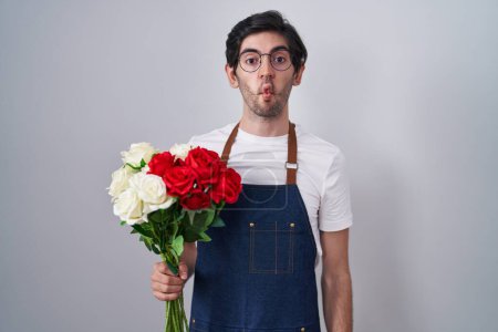 Photo for Young hispanic man holding bouquet of white and red roses making fish face with lips, crazy and comical gesture. funny expression. - Royalty Free Image