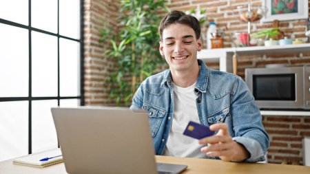 Téléchargez les photos : Jeune homme hispanique shopping avec ordinateur portable et carte de crédit assis sur la table à la salle à manger - en image libre de droit